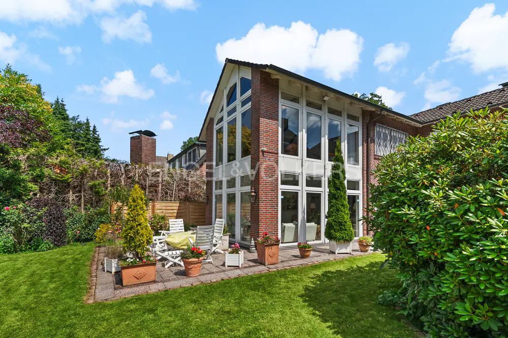 Exterior view of a brick house with large windows, a patio with white chairs, and a green lawn under a blue sky.