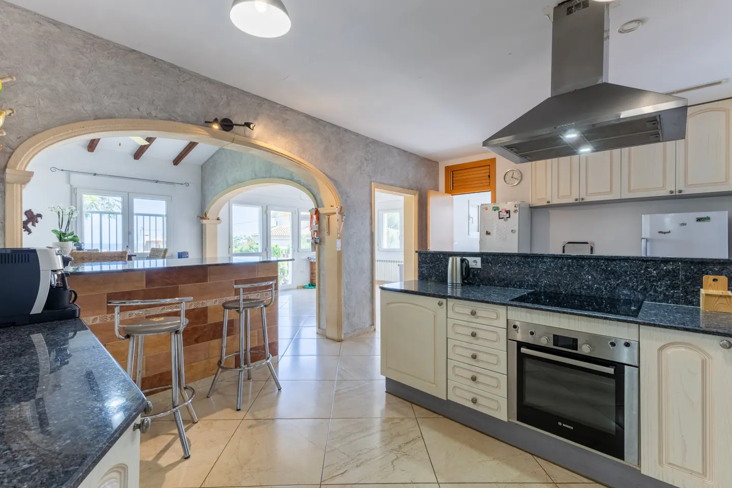A bright kitchen with cream cabinets, black countertops, and stainless steel appliances. A tiled bar with stools separates the kitchen from a sunlit room.