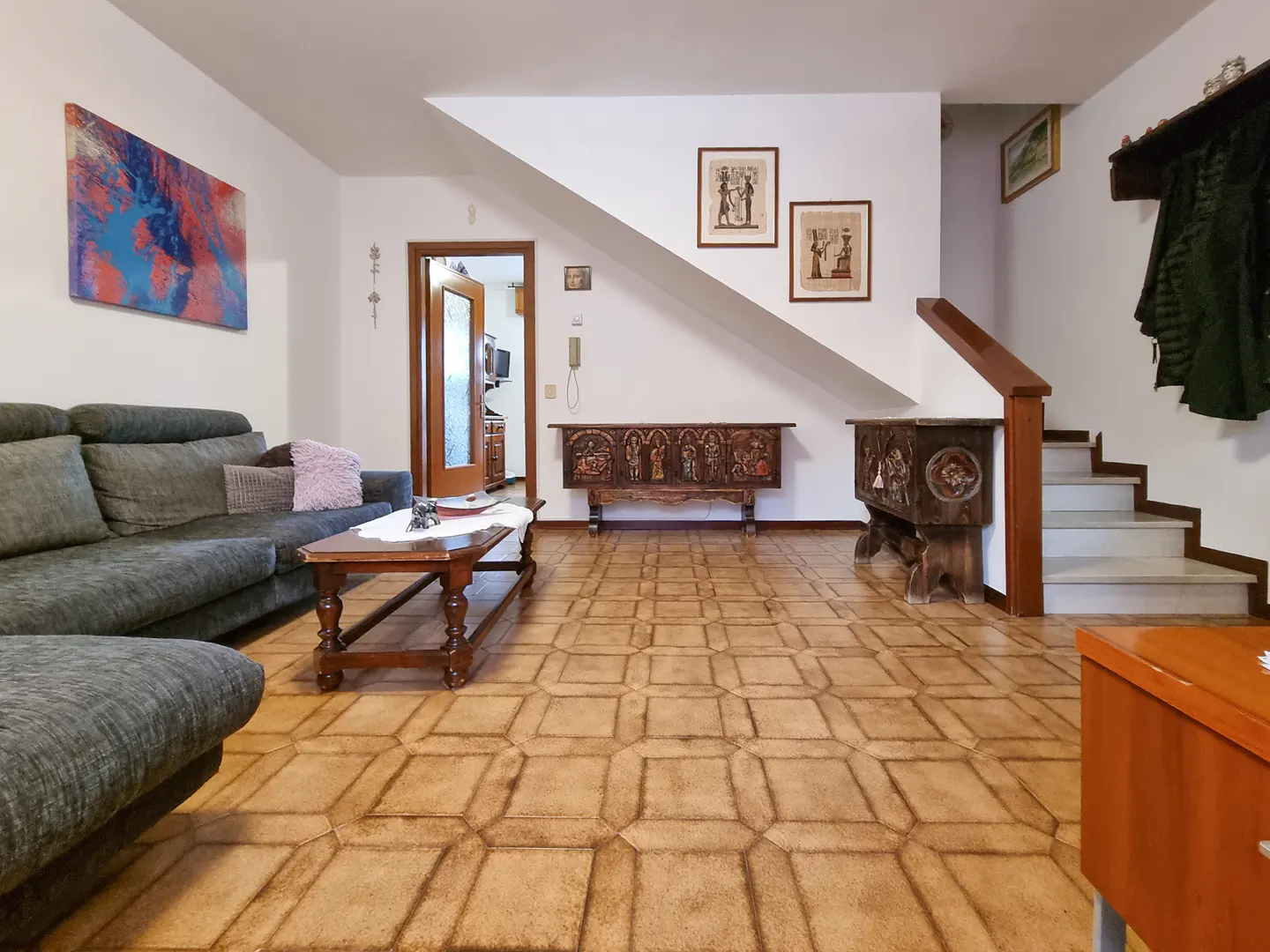 A living room with a gray sofa, wooden coffee table, and patterned tile floor. Stairs lead to the upper level. Carved wooden chests are against the wall.