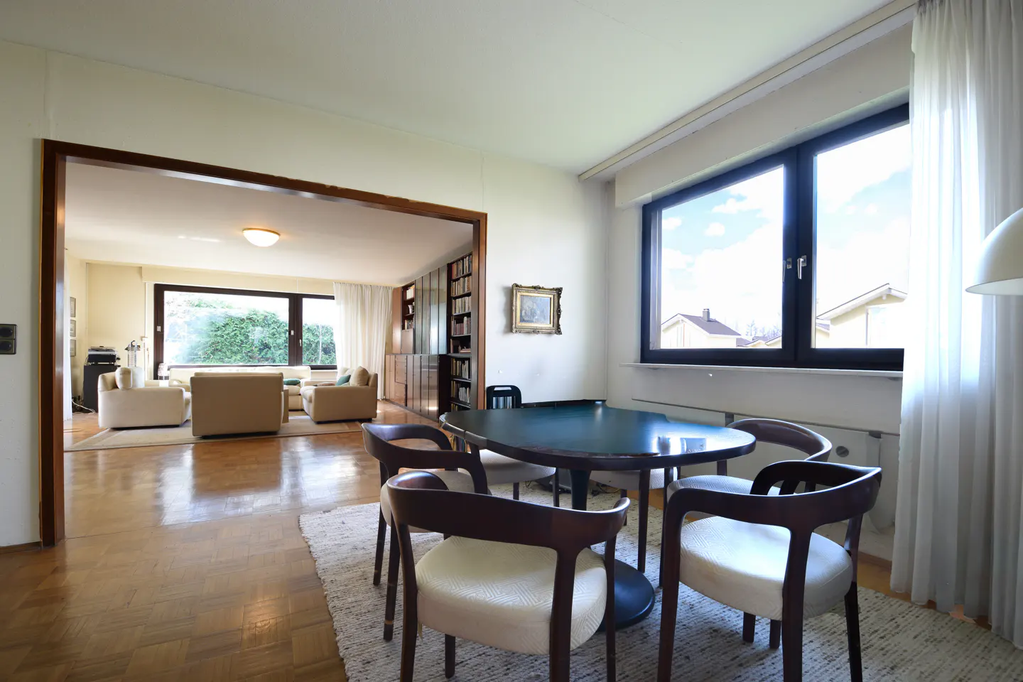 A bright, open-concept living and dining area with wood floors, a dark green table, and large windows.