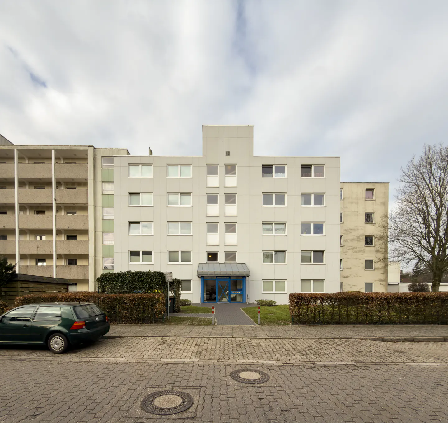 Exterior of a light gray apartment building with a blue entrance and a green car parked nearby.
