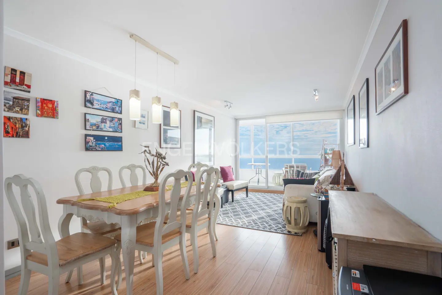 Bright dining area with a wooden table, six chairs, and a living room beyond with a view of the ocean.