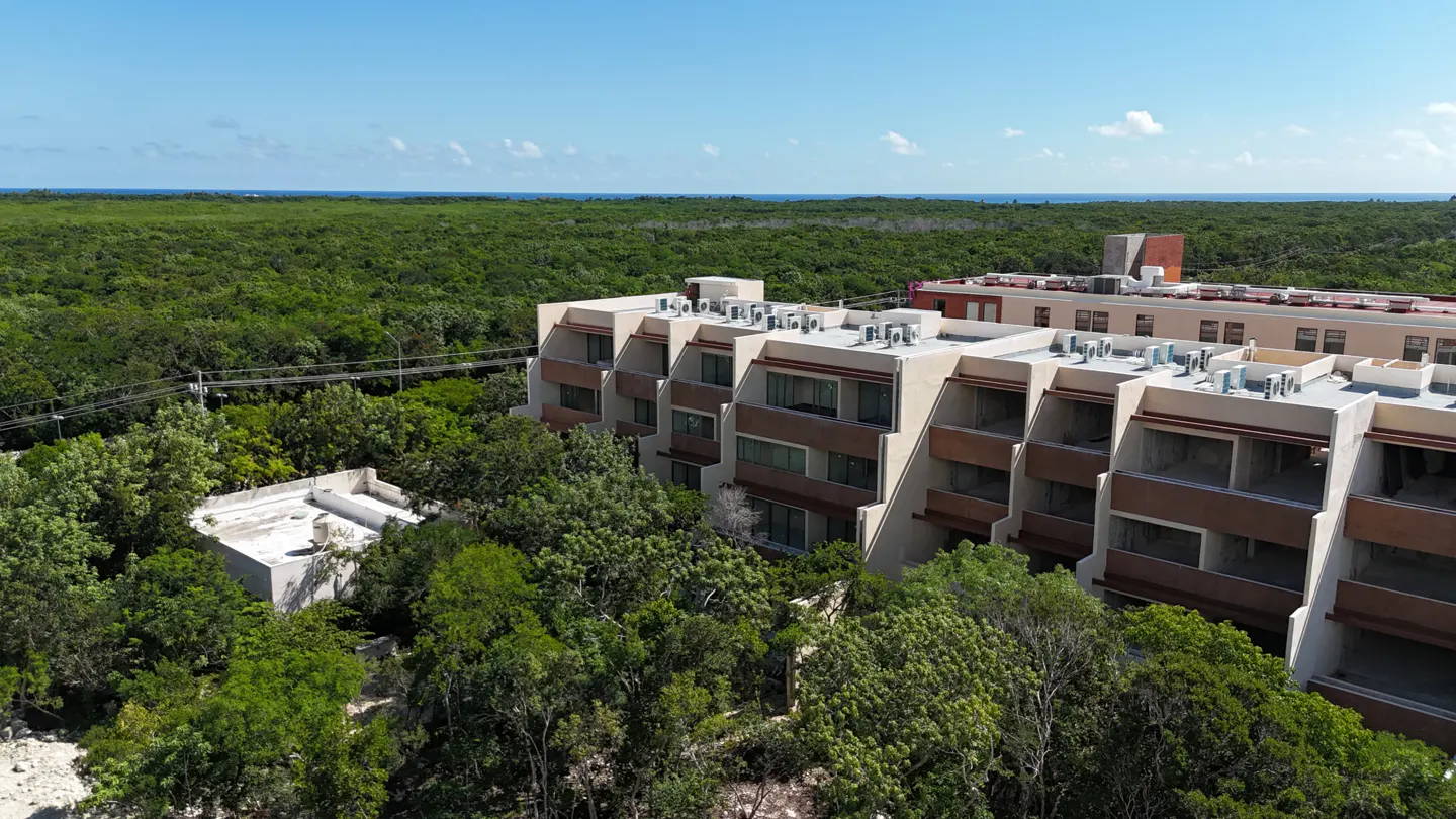 Aerial view of a multi-story beige and brown apartment building surrounded by dense green forest near the ocean.