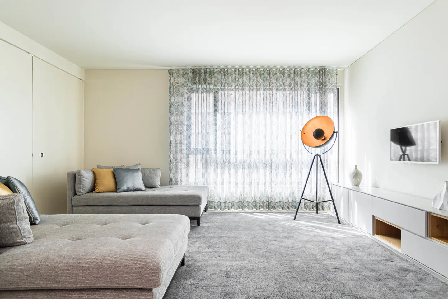 Bright living room with gray sofas, pillows, and carpet. A large window with patterned curtains and a tripod floor lamp provide light. A white console table and TV are on the right.
