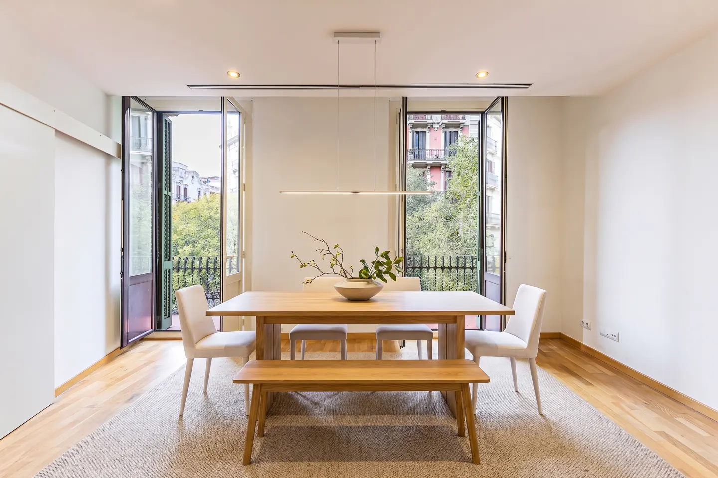 Modern dining room with a wooden table, bench, and white chairs on a rug, framed by open balcony doors overlooking trees.