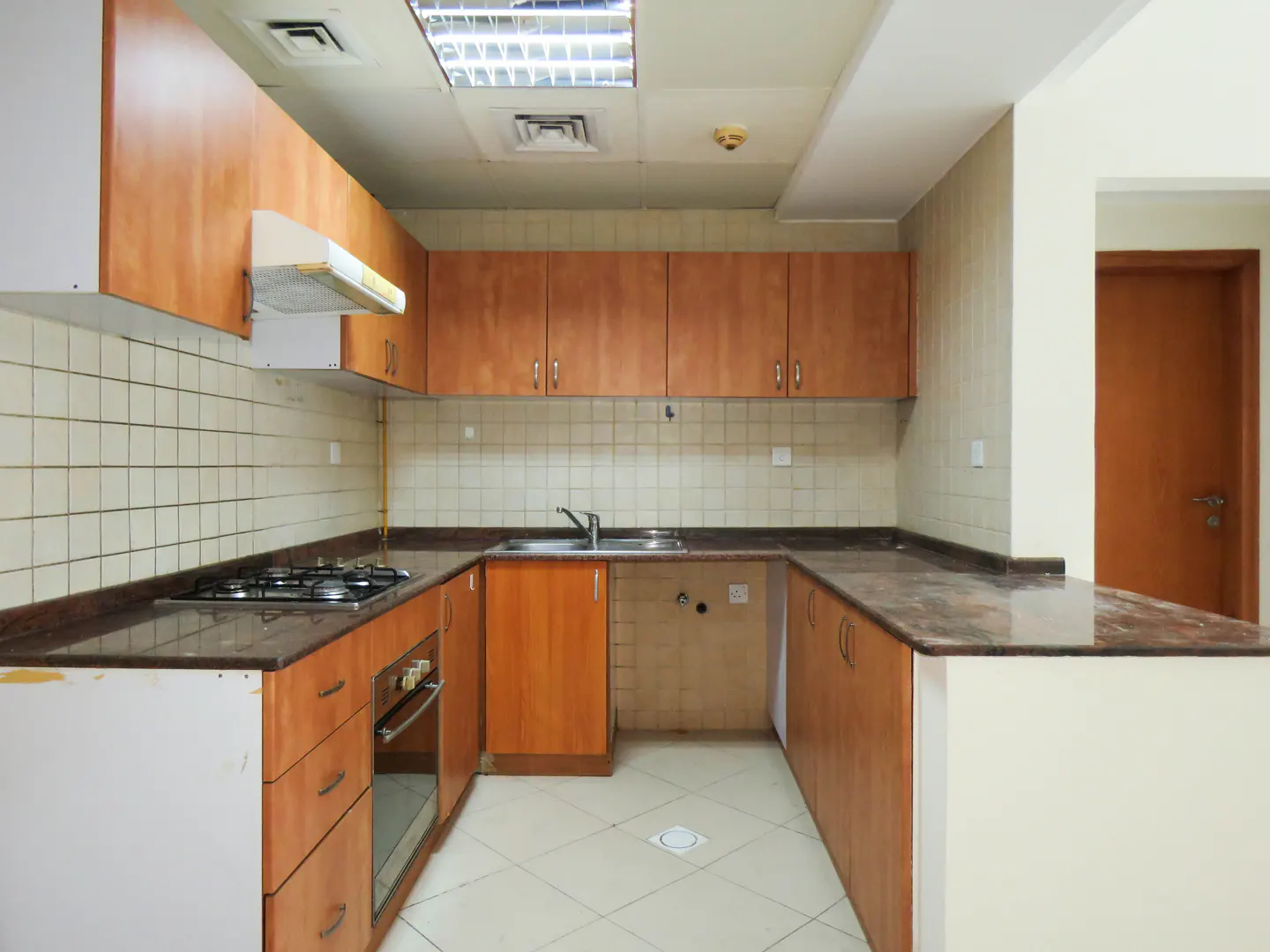 A kitchen with wood cabinets, a gas stove, and a marble countertop. The floor is tiled, and there is a door visible in the background.