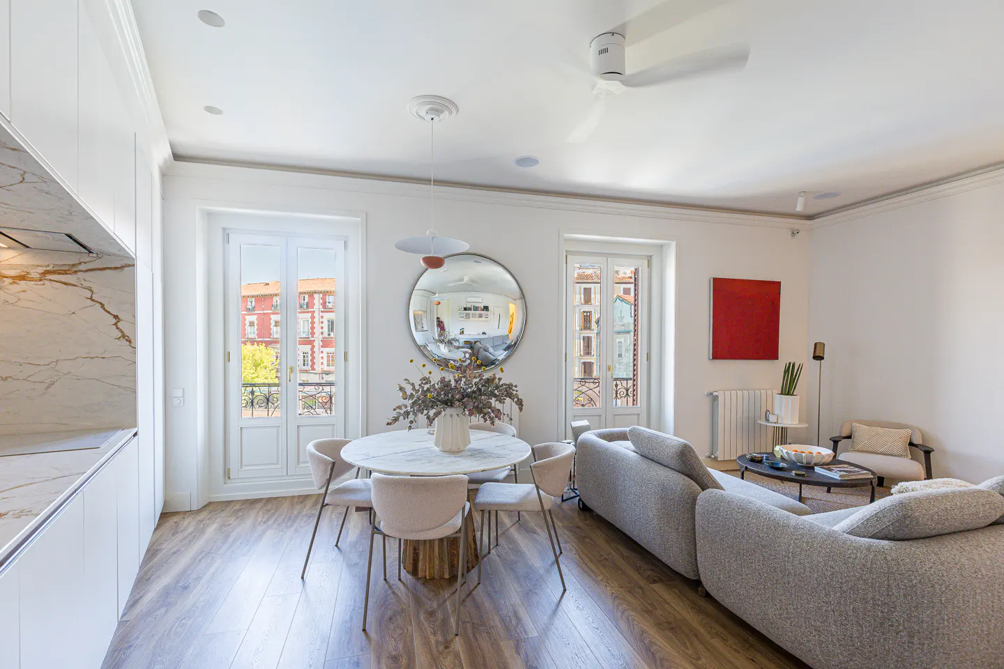 Bright, modern living space with wood floors, white walls, and a gray sofa. A round marble table with chairs sits near balcony doors.
