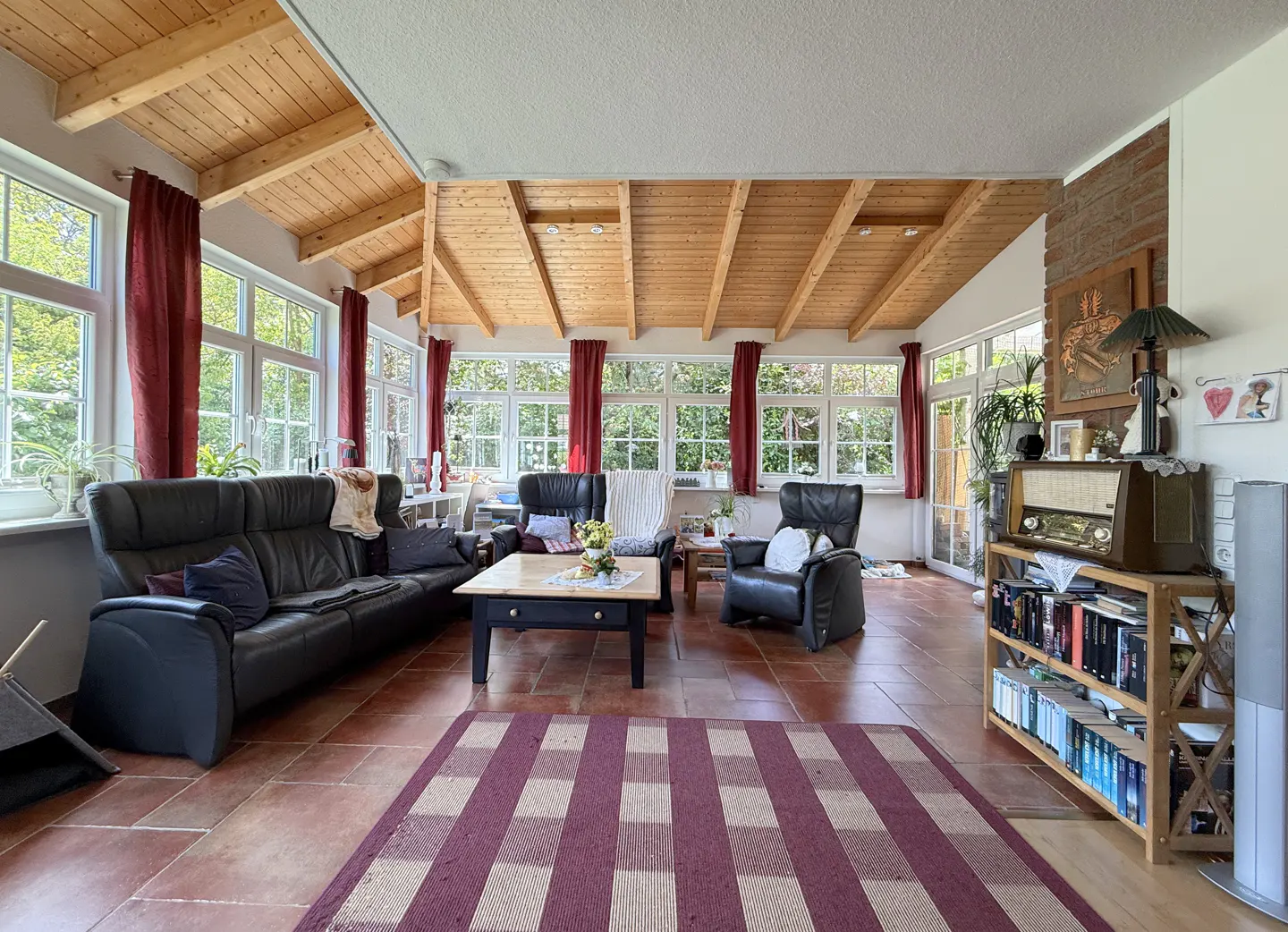 Sunroom with wood-beamed ceiling, black leather sofa and chairs, red curtains, tiled floor, and a vintage radio on a shelf.