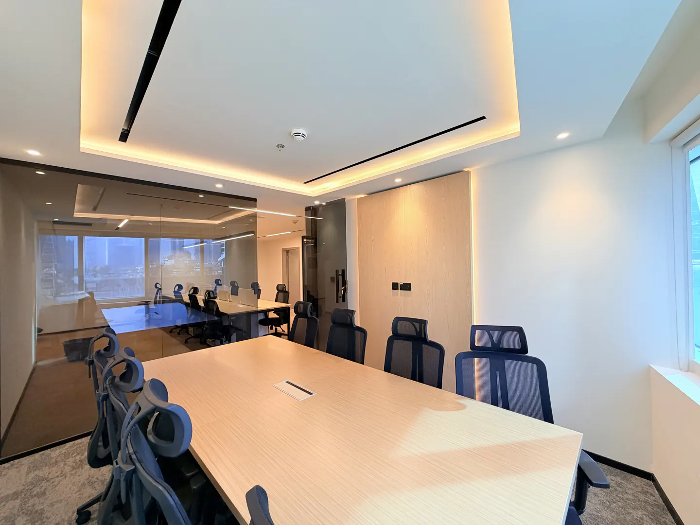 A modern conference room with a long, light wood table and blue mesh chairs. A glass-walled office is visible in the background.