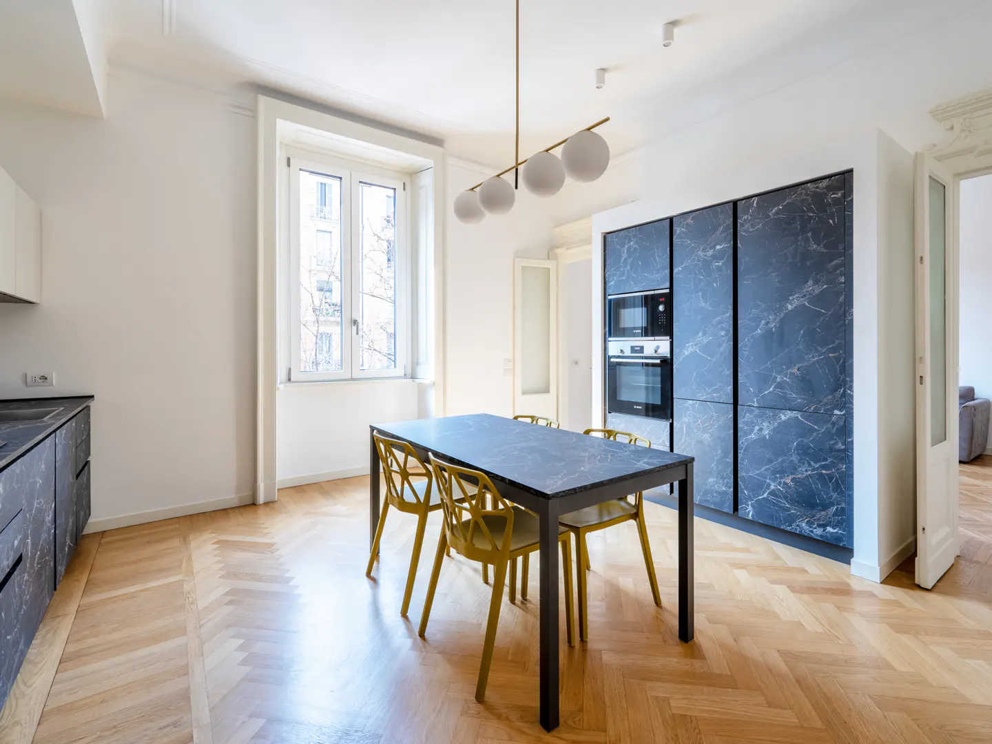 Bright kitchen with herringbone wood floors, a black marble table with yellow chairs, and a modern light fixture.