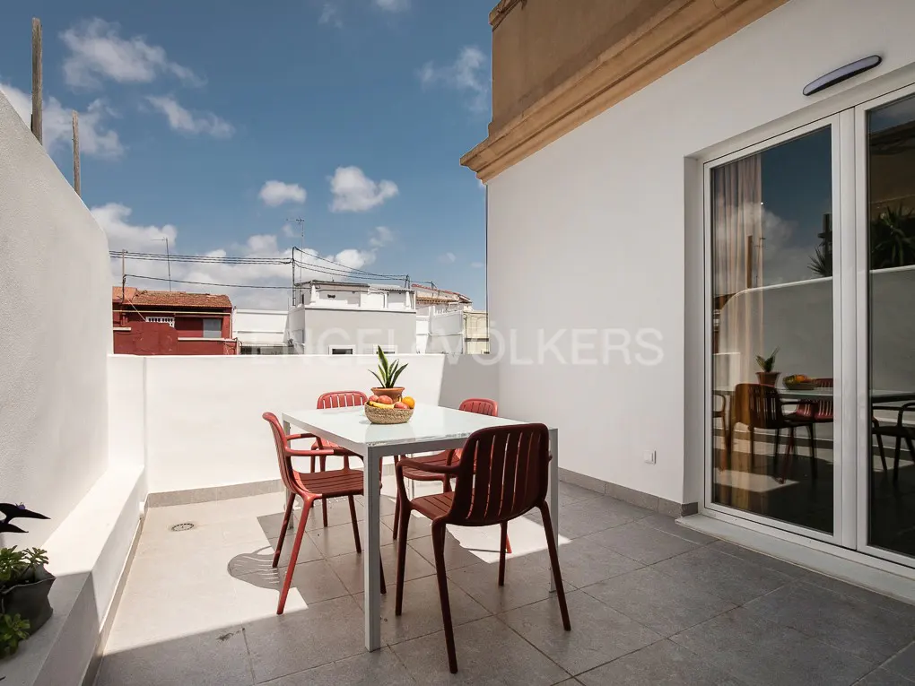Outdoor patio with white walls, gray tile floor, and a white table with four red chairs. A sliding glass door reflects the table setting. Blue sky.