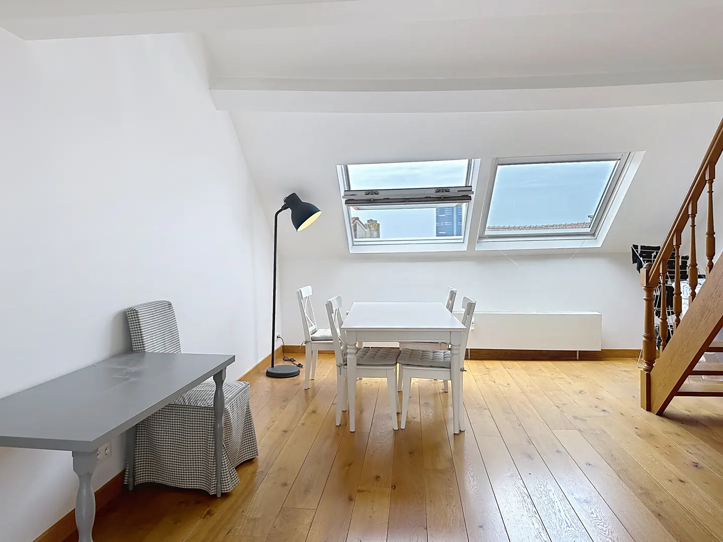 Bright attic room with wood floors, white walls, and skylights. A white table and chairs sit near a black floor lamp. A gray desk is on the left.