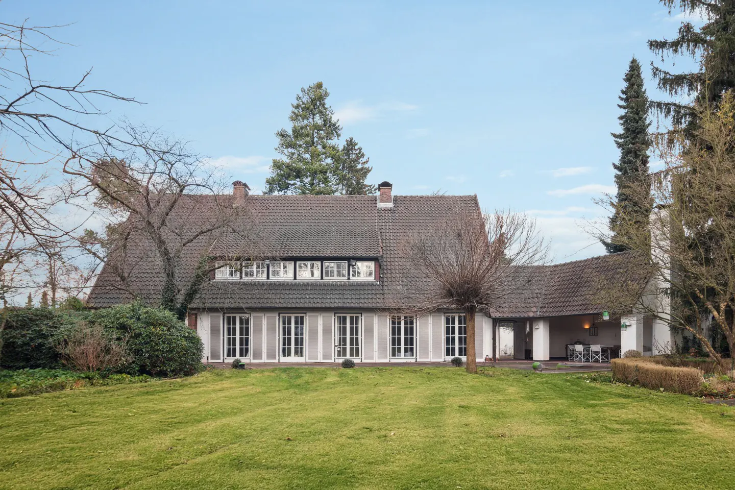 Exterior view of a large, two-story house with a gray roof, white shutters, and a green lawn. Trees surround the property.