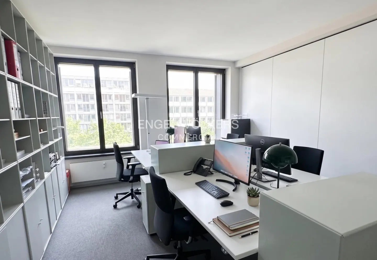 Bright office space with white desks, black chairs, and large windows. A grey shelving unit is on the left.