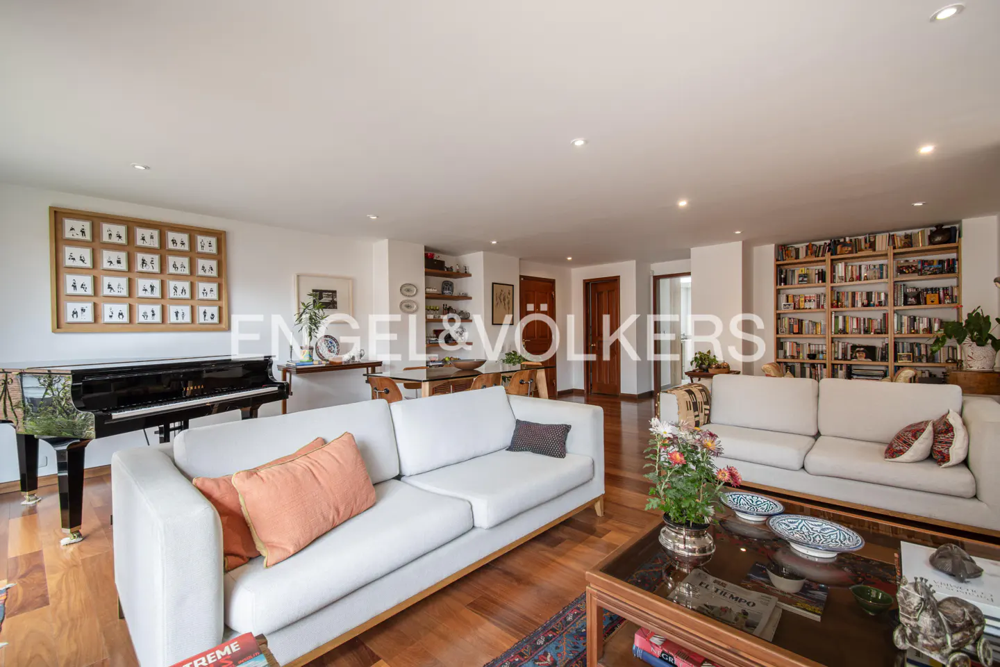 A wide shot of a living room with white sofas, a piano, and a bookshelf.