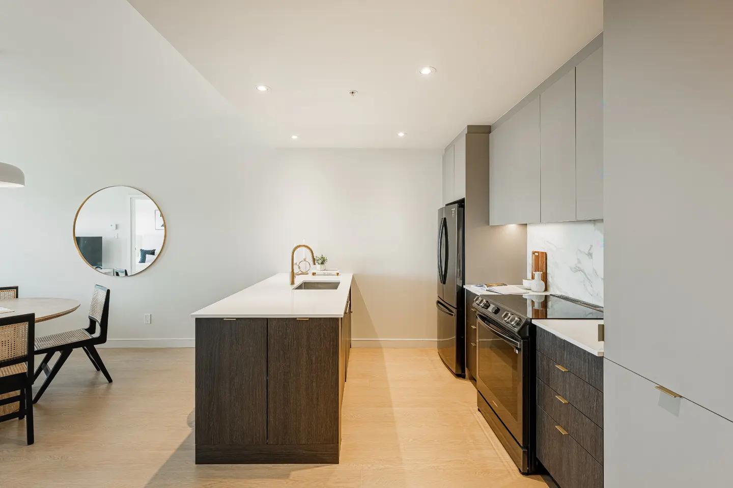 A modern kitchen with a dark wood island, white countertops, and stainless steel appliances. A round mirror hangs on the wall near a dining table.