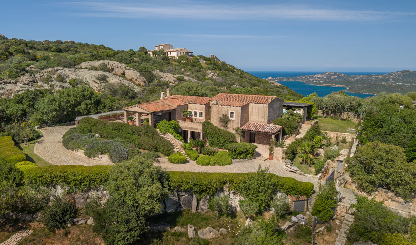 Aerial view of a tan house with a red tile roof, surrounded by green trees and bushes, with a blue ocean in the background.