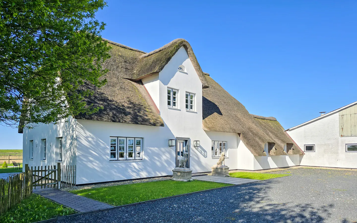 Exterior of a white house with a thatched roof, statues, and a gravel driveway under a clear blue sky.
