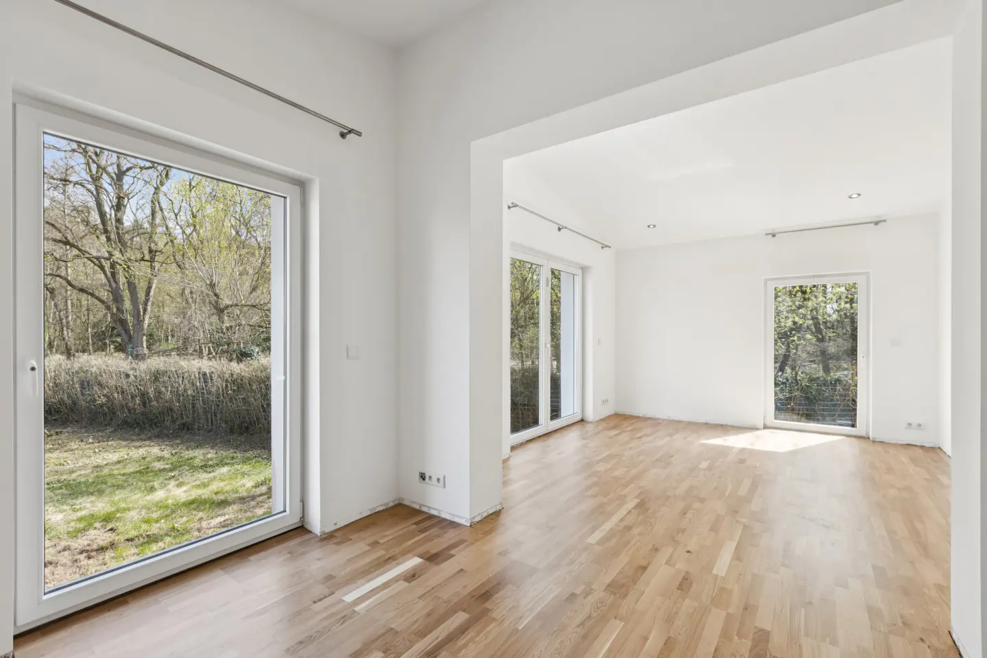 Bright, empty room with hardwood floors and white walls. Large windows offer views of trees and greenery.