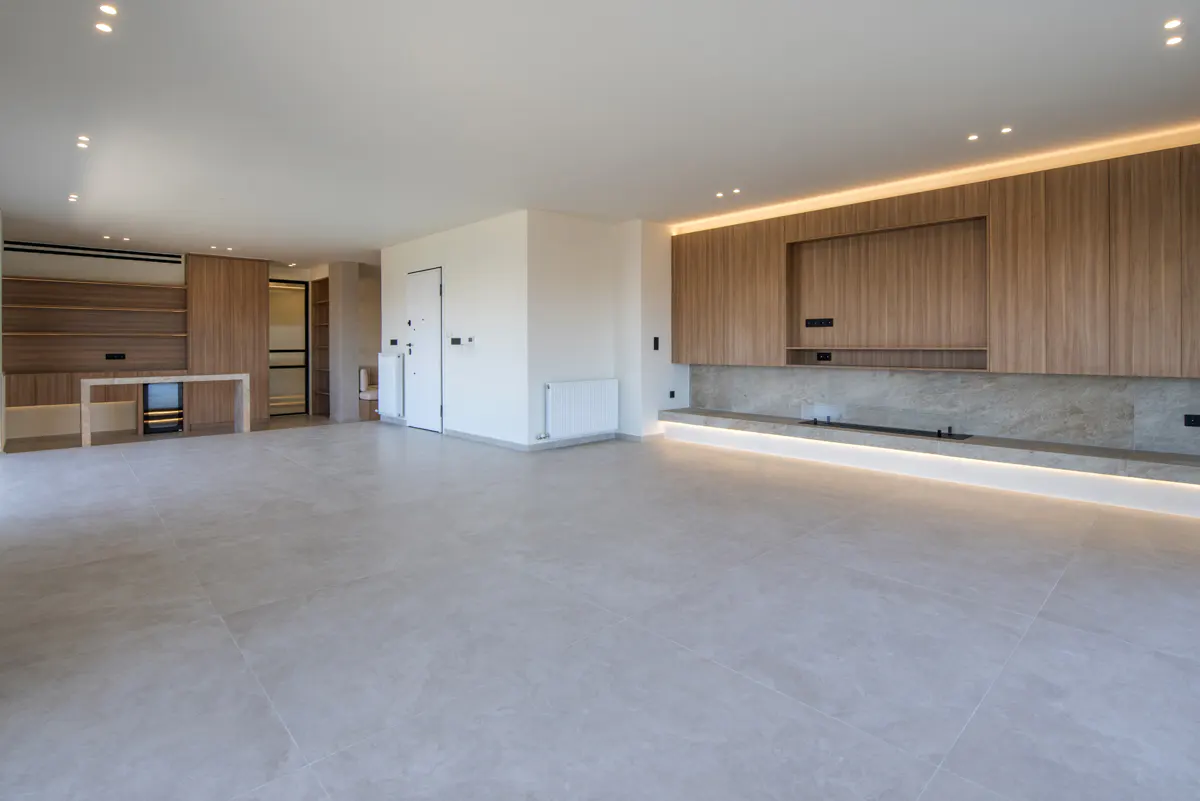 Bright, empty living room with light gray tile floor, wood cabinets, and white walls. Recessed lighting and under-cabinet lights add warmth.