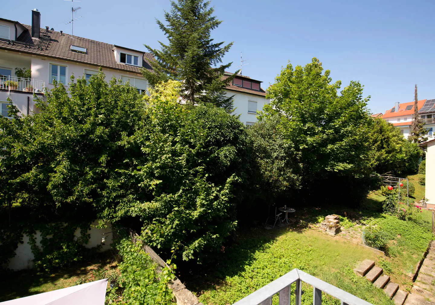 View from a balcony overlooking a lush green backyard with trees, grass, and a small table and chairs. Buildings are visible in the background.