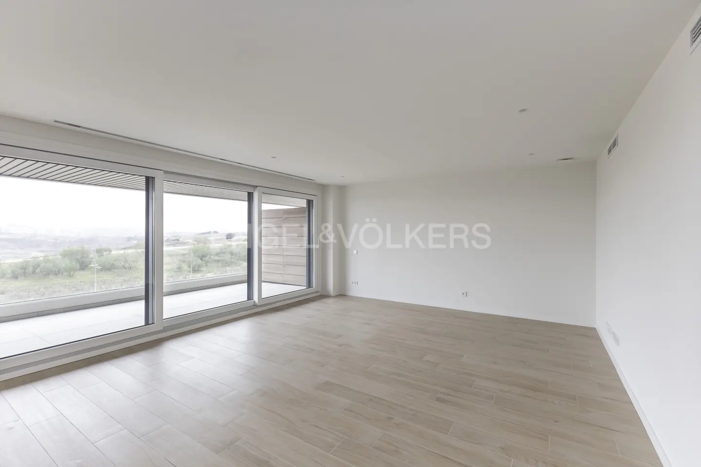 Bright, empty room with light wood floors, white walls, and large windows overlooking a landscape. Engel & Völkers logo on the wall.