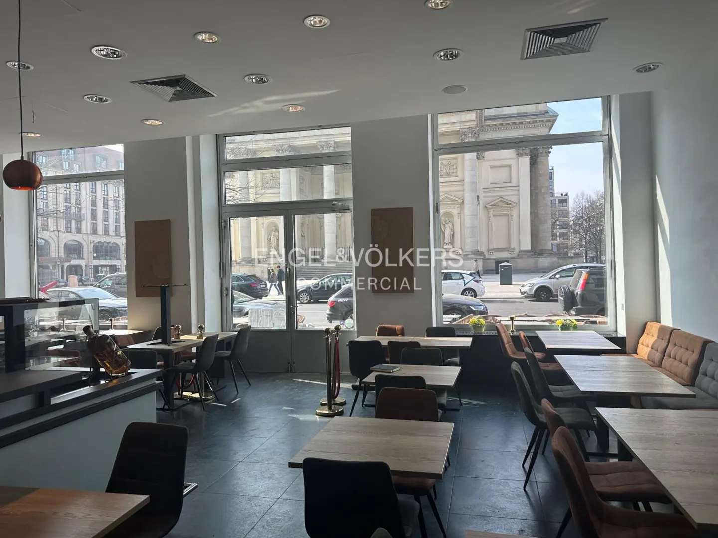 Interior view of a bright restaurant with tables and chairs, large windows overlooking a city street with cars and buildings.