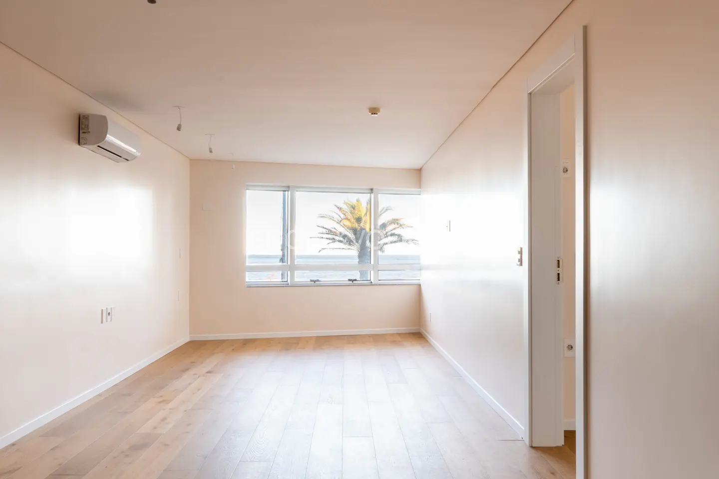 Bright, empty room with light wood floors, beige walls, and a large window showing a palm tree and ocean view. An air conditioner is mounted on the wall.