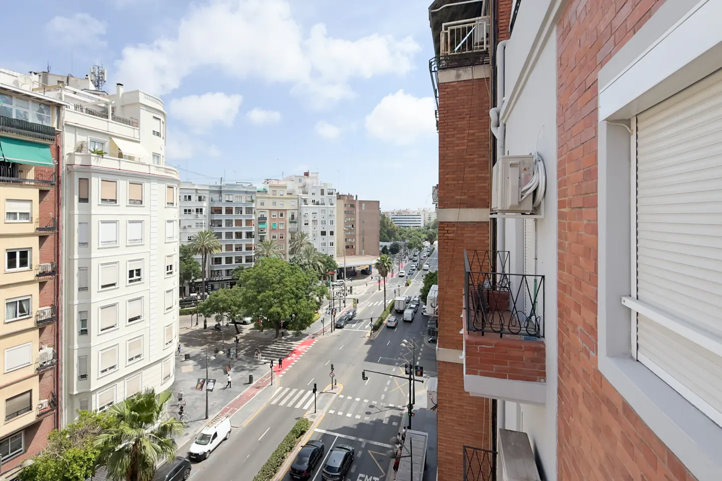 View from a balcony overlooking a city street with cars, crosswalks, and buildings under a blue sky with clouds.