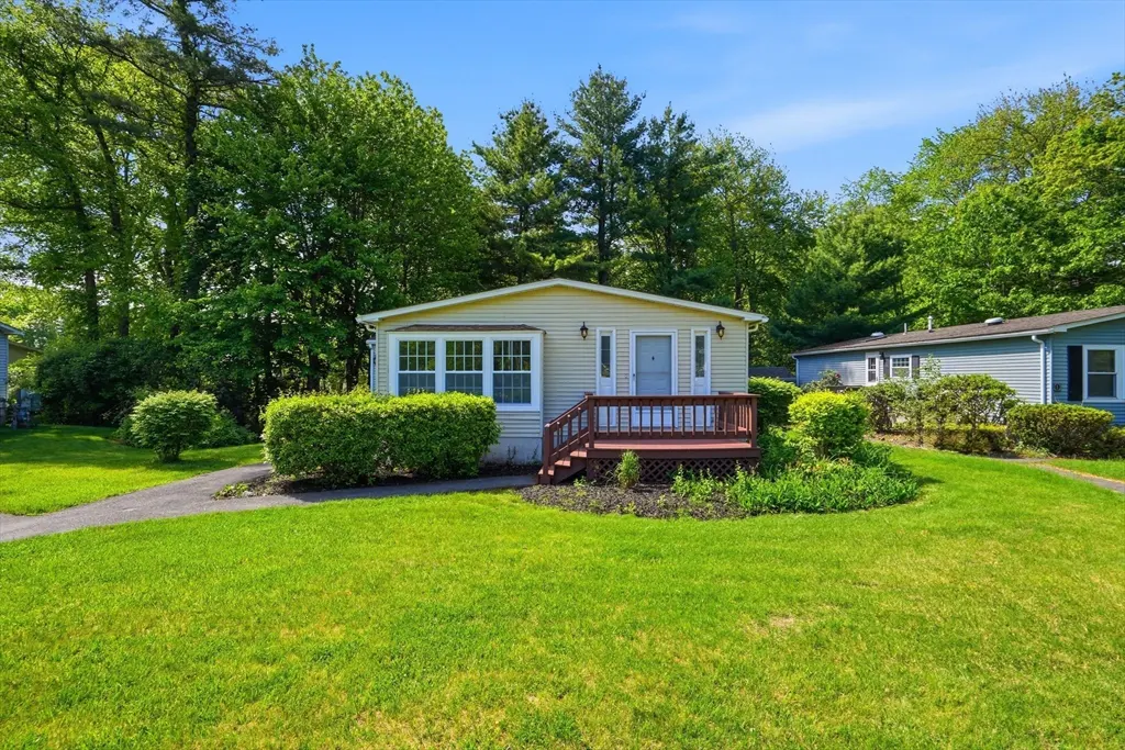 A small, beige house with a brown porch and green lawn sits in front of a lush green forest.
