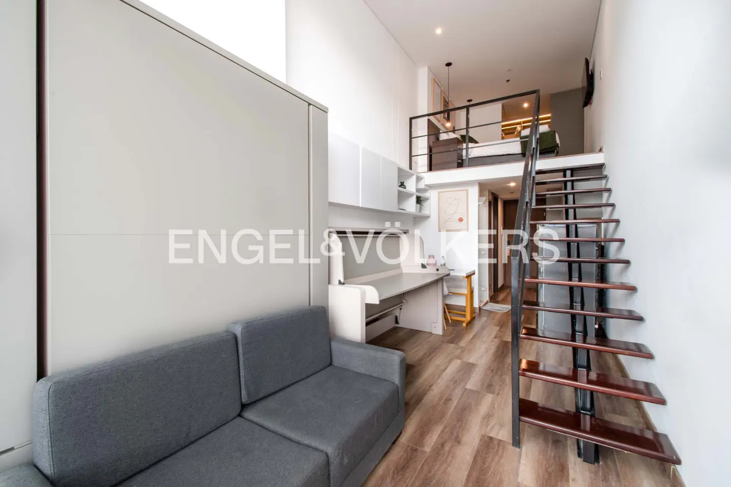 Modern apartment interior with gray sofa, desk, and stairs leading to a loft bedroom. Wood floors and white walls.