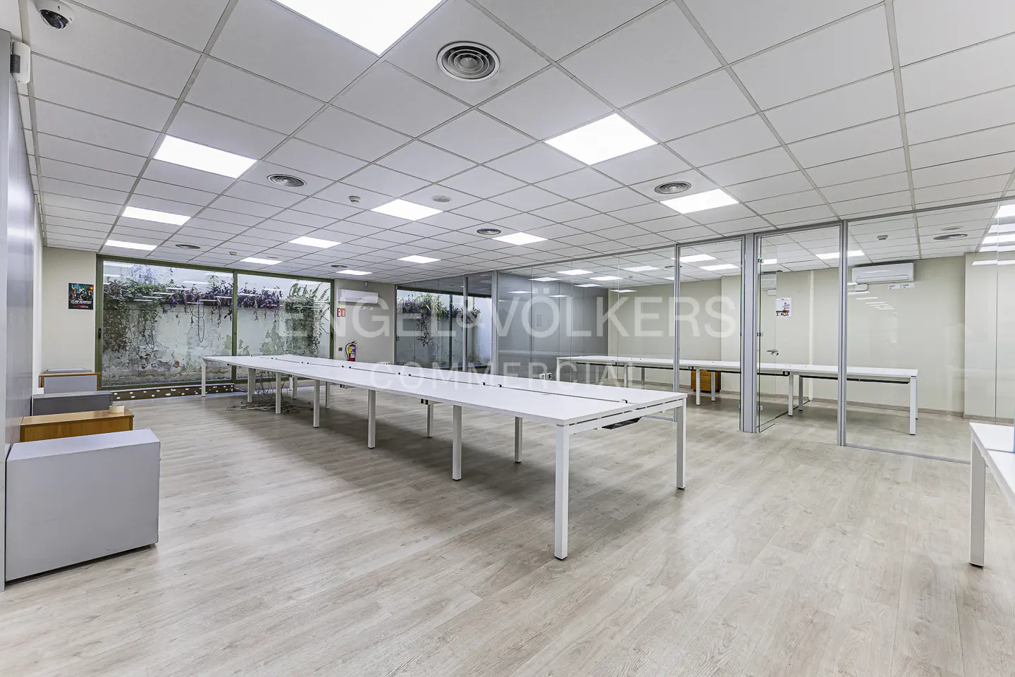 Bright office space with white tables, light wood floors, and a glass-walled room. Windows overlook greenery.
