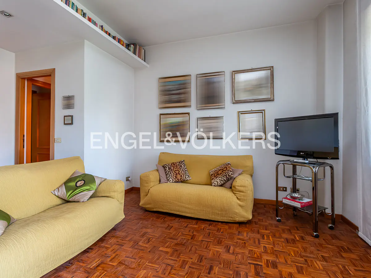 Living room with two yellow sofas, patterned pillows, wood floors, and a TV on a metal stand. Artwork hangs on the white wall.
