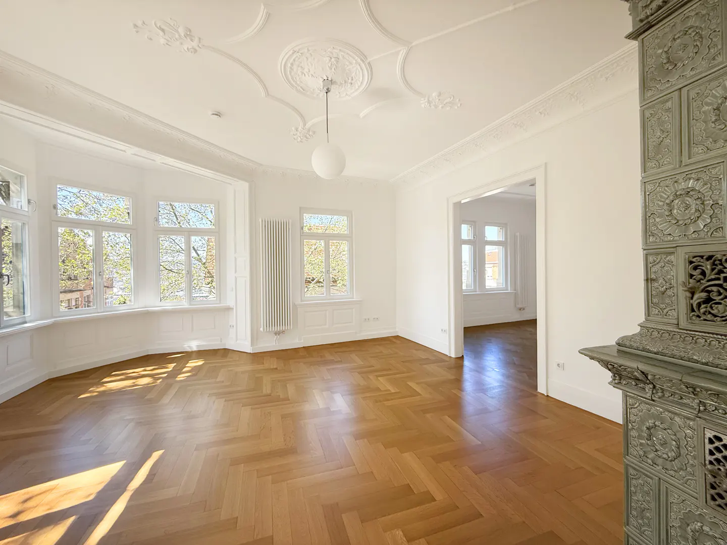 Bright, empty room with herringbone wood floors, white walls, and ornate ceiling. Windows let in natural light. A tiled stove stands in the corner.