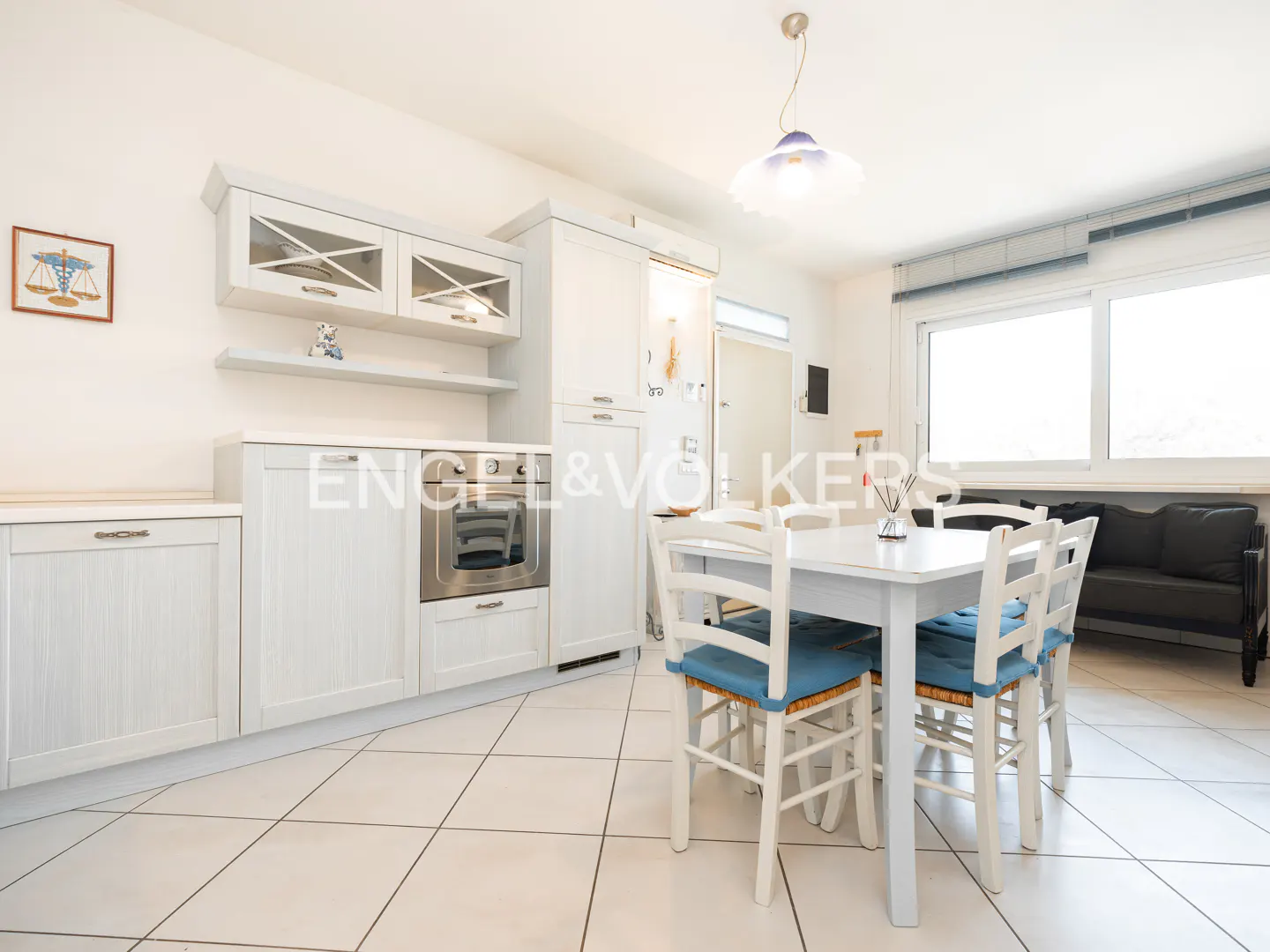 Bright kitchen with white cabinets, stainless steel oven, and a white table with blue-cushioned chairs. A black sofa sits near a large window.