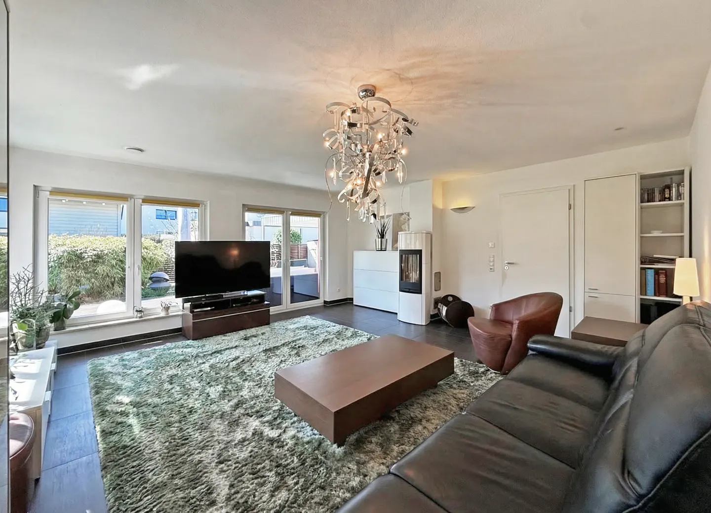 Bright living room with a black leather sofa, brown coffee table, and a green shag rug. A modern chandelier hangs from the ceiling. Large windows offer a view of the outdoors.