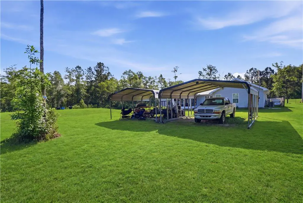 A light blue house with a truck parked under a metal carport on a large green lawn.