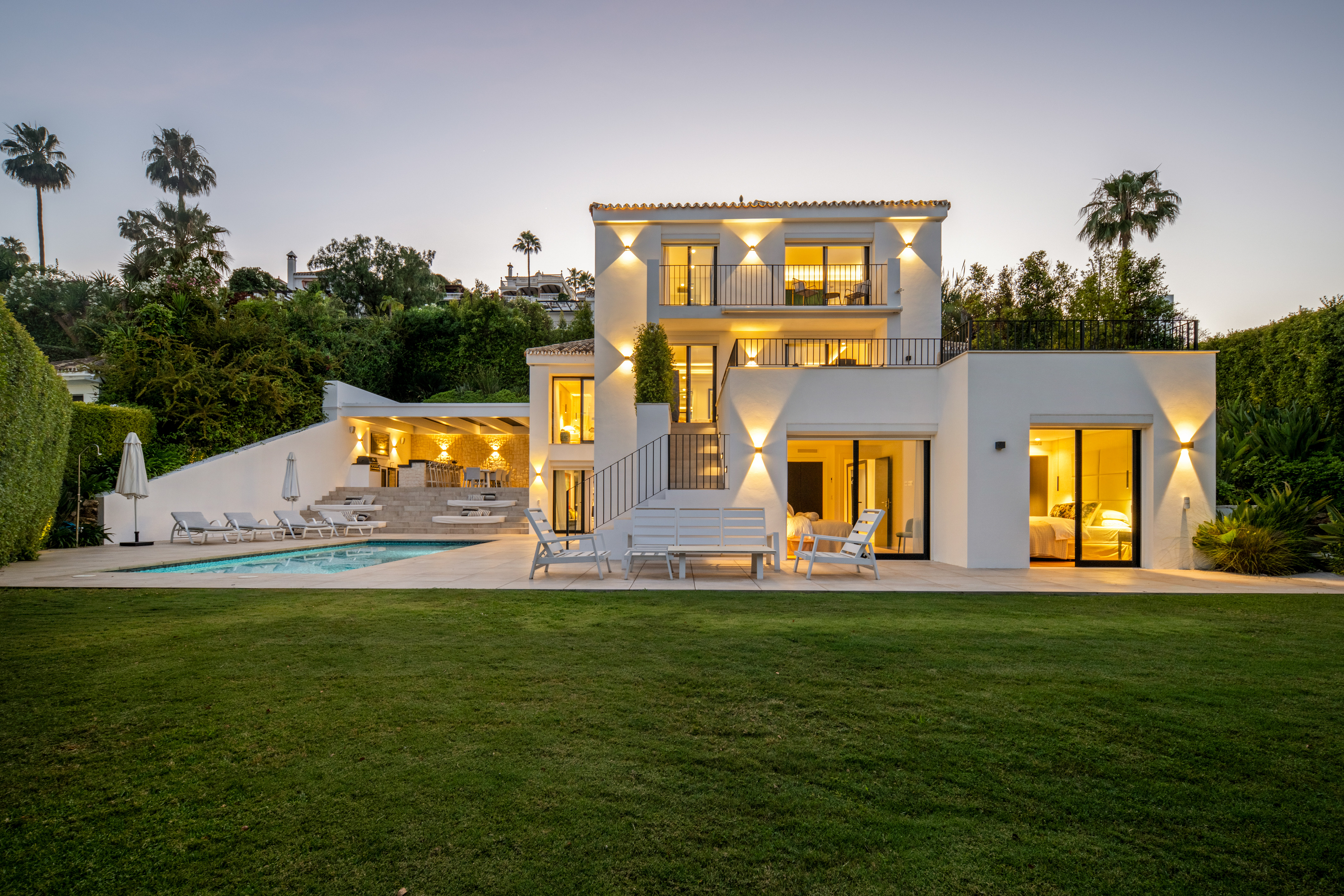 Modern white villa at dusk with a swimming pool, outdoor lounge area, illuminated balconies, and a lush green lawn.