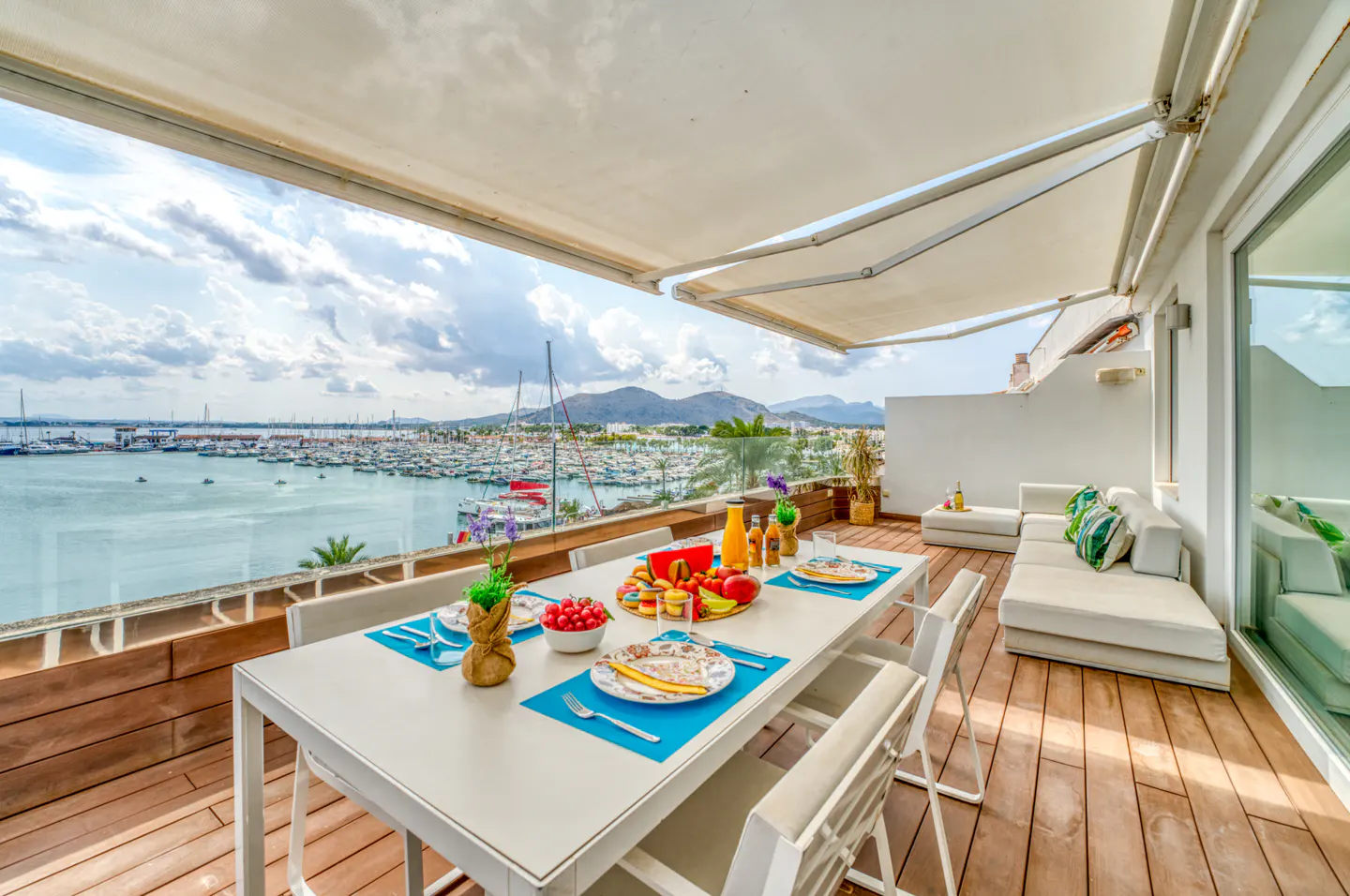 Outdoor patio with a white table set for a meal, overlooking a harbor filled with boats and a mountain range in the distance.