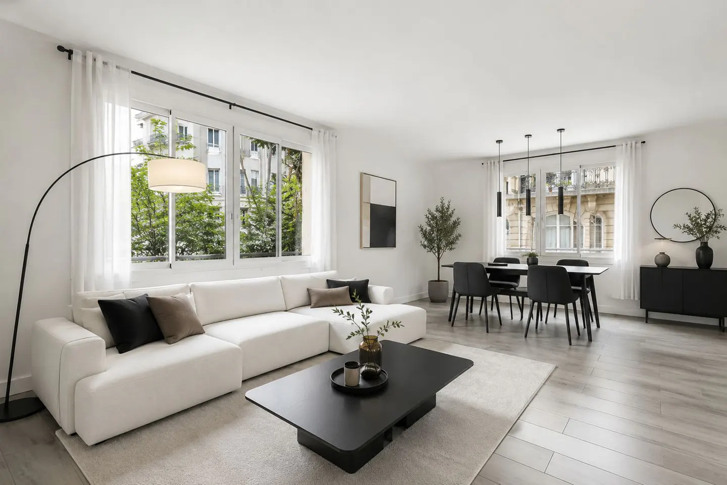 Bright, modern living room with white walls, large windows, and light wood floors. A white sectional sofa faces a black coffee table and dining set.