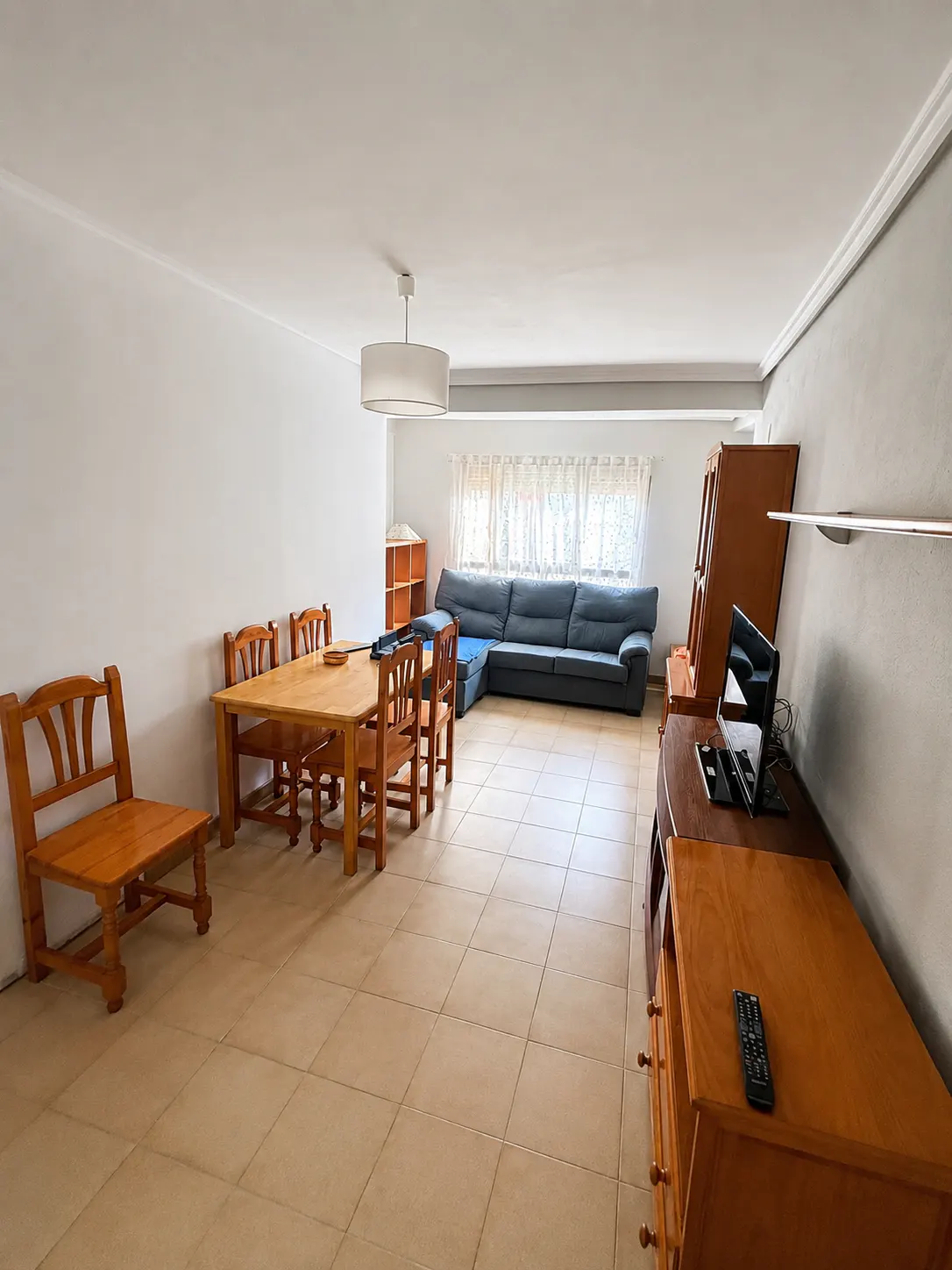 A bright living room with a wooden dining table, chairs, a blue sofa, and a TV stand.
