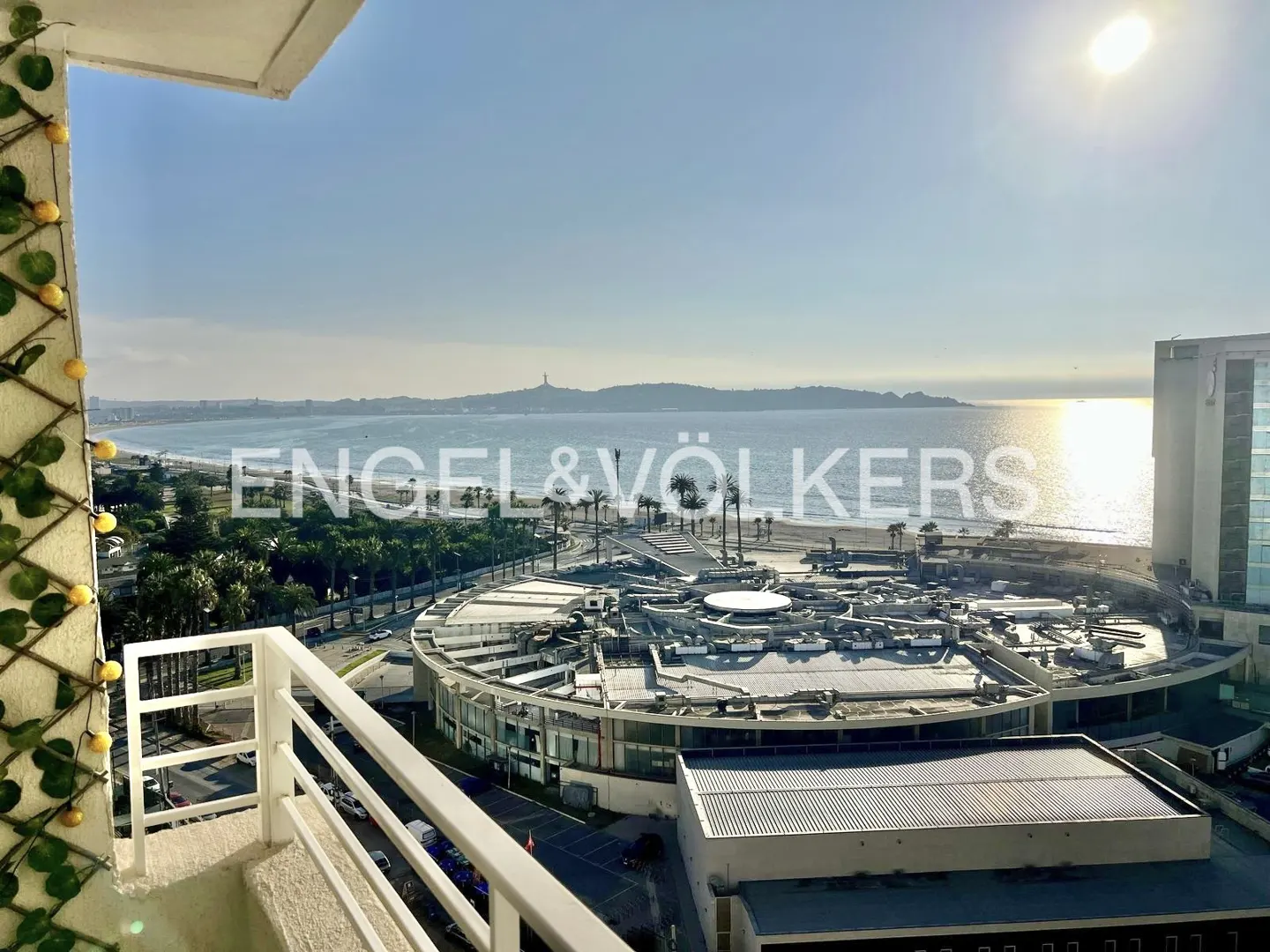 Balcony view of a sunny beach and city. White railings frame the scene, with a decorative vine on the left. Ocean and buildings stretch to the horizon.