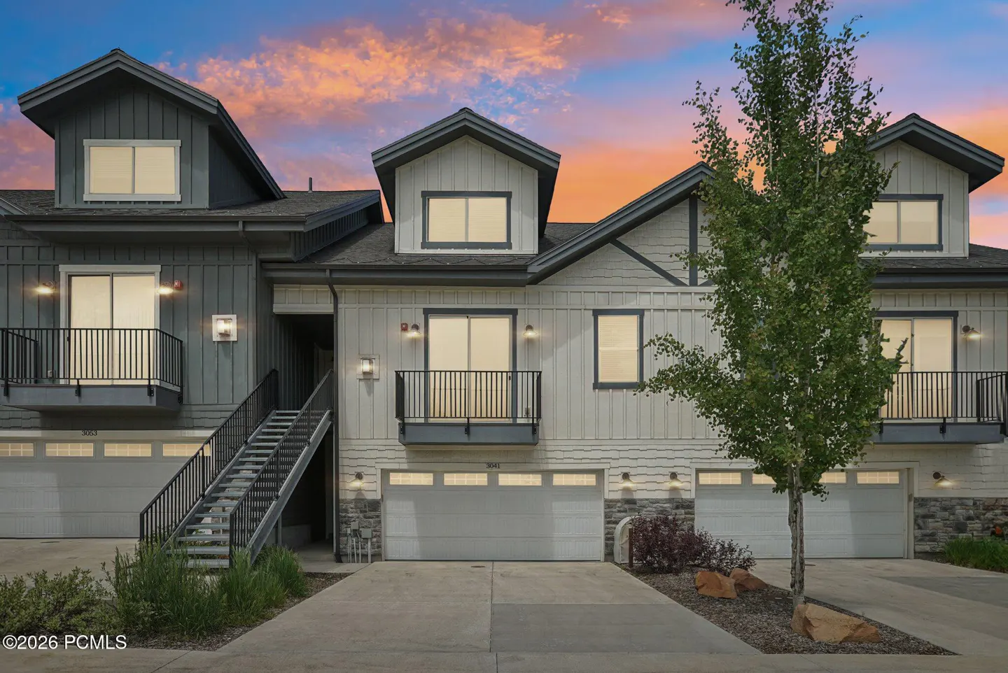 Exterior view of a three-unit townhouse with gray and white siding, black trim, and individual garages at dusk.