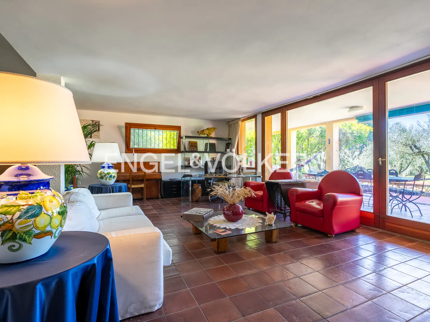 Bright living room with red chairs, white sofa, and terracotta tile floor. Large windows overlook a patio and garden.