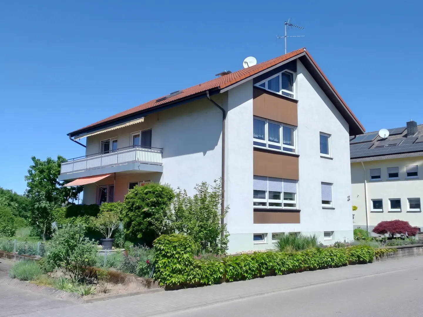 Two-story house with white walls, brown accents, and a red tile roof under a clear blue sky. A balcony is on the left side.