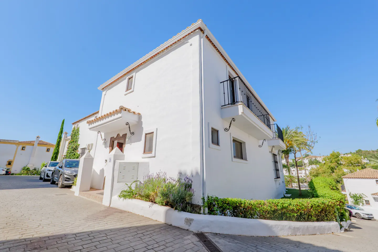 A two-story white Mediterranean-style house with a balcony, set on a paved street under a clear blue sky.