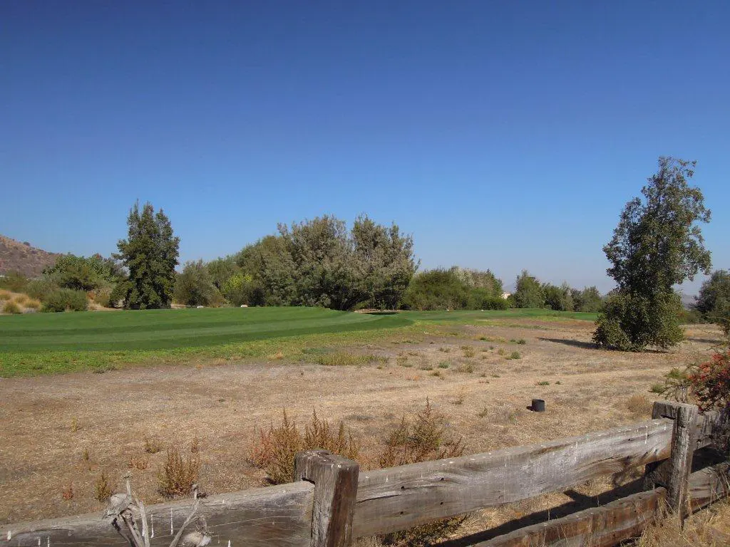 A sunny view of a golf course with a weathered wooden fence in the foreground. Green grass and trees are visible under a clear blue sky.