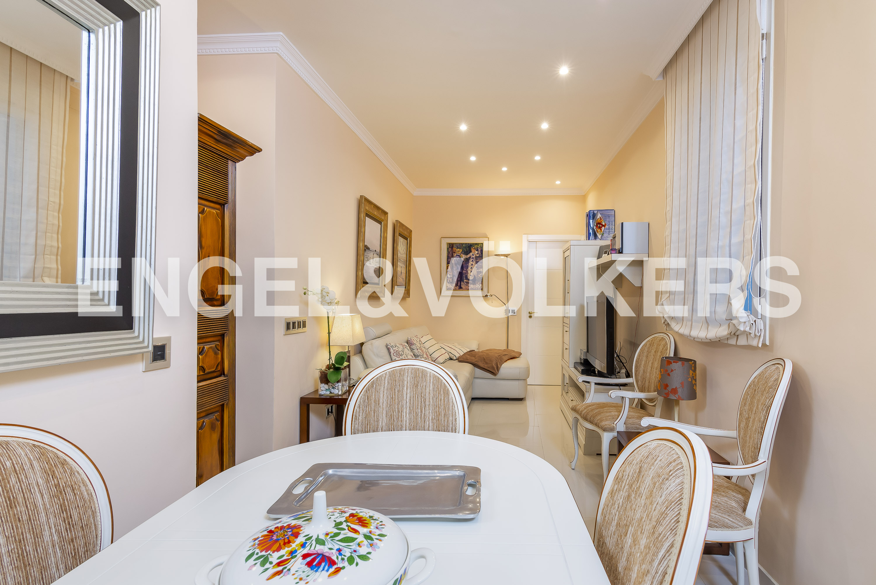 Dining area with a white oval table, floral tureen, and upholstered chairs, leading to a cozy living room with a sofa.