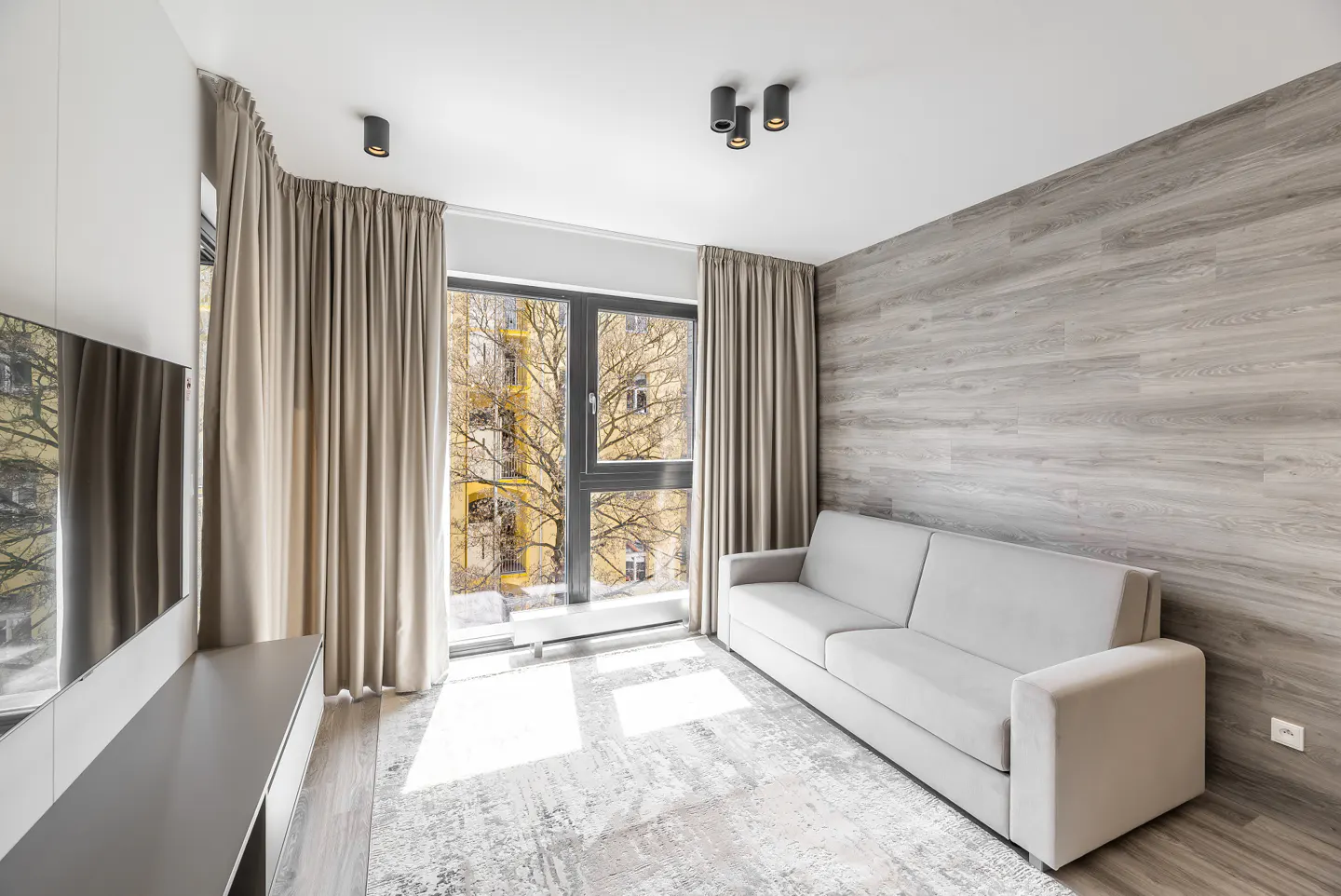 Living room with a gray sofa, wood-look accent wall, and large window with beige curtains. A gray rug covers the floor.