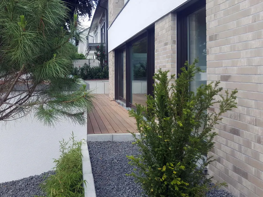 Exterior view of a modern home with a brick facade, dark-framed windows, and a wooden deck surrounded by greenery and gray gravel.